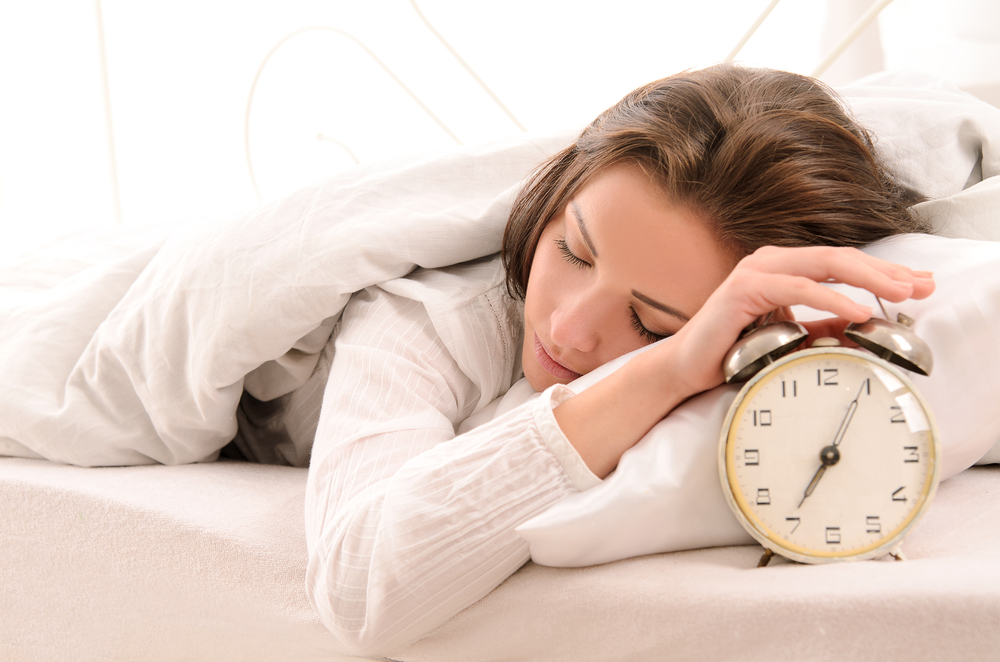 woman with brown hair sleeping in a white bed with her hand on her alarm clock